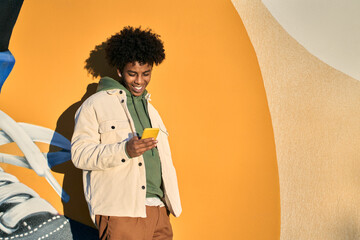 Happy smiling cool gen z young African American ethnic stylish hipster guy model standing at yellow city urban wall using cell phone mobile device, looking at smartphone, holding cellphone. - obrazy, fototapety, plakaty