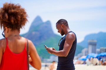 waist up portrait young black brazilian man standing at the beach looking at a cell phone with two...
