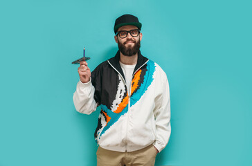 Fashionable bright man with a beard in a vintage sports jacket, cap and glasses with an audio cassette in retro style isolated on a blue studio background. Old school