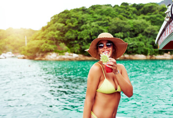 portrait young brazilian woman in swimsuit on a boat drinking juice happy with water and vegetation...