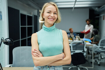 Smiling blonde Caucasian business woman looking at camera standing with arms crossed in office. Executive CEO professional manager posing for business portrait.