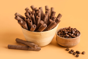 Bowls with delicious chocolate wafer rolls and coffee beans on orange background