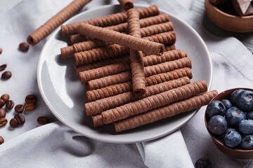 Plate with delicious chocolate wafer rolls and blueberries on grey wooden background