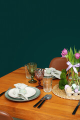 Festive dining table with painted eggs and tulip flowers served for Easter celebration near green wall
