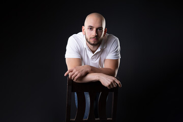 turkish student man with bald and white shirt lean on chair back with arms crossed while look at camera  with neutral and serious face expression