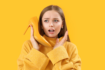 Fashionable surprised woman in knitted sweater holding high-heeled shoe on yellow background