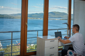 A man is working at a computer in an office with large panoramic windows overlooking the sea.