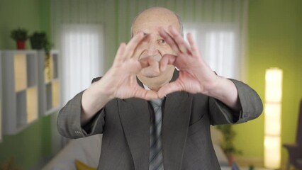 Cute and romantic old man making heart sign at camera.
Sympathetic naive old man looking at camera at home making heart sign, feeding love.
