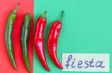 Fotobehang Four hot green and red chili peppers with the inscription on paper: fiesta and copy-space lie on a red-green. © Nataliya