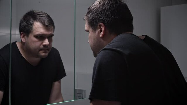 Upset lonely man suffering from depression looking in reflection at bathroom mirror. Concept of depression, suicide, stress, mental illness, loneliness and frustration