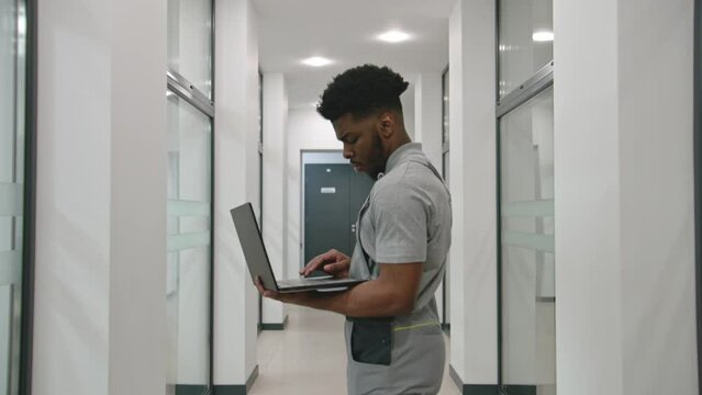 African American installer in uniform stands in hallway and sets up security cameras using program on laptop. CCTV cameras installation in coworking office. Concept of surveillance system. Dolly zoom.
