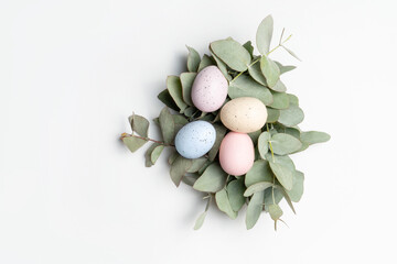 Pastel coloured easter eggs, in a rustic nest made of eucalyptus leaves, on a white background, photographed from above.