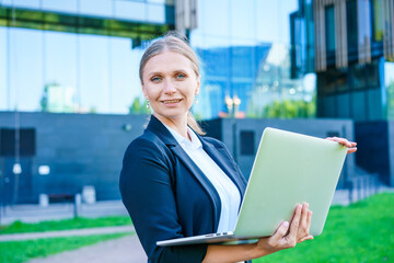 Portrait confident successful young woman, formally dressed, business woman, standing with a laptop outdoors against the backdrop of a business center