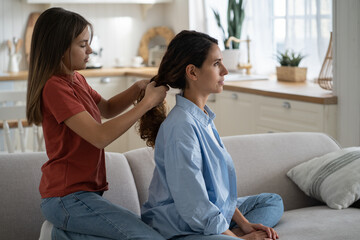Family time. Pre-teen girl daughter braiding mothers long wavy hair, selective focus. Mom and child enjoying time with each other on weekend at home. Child making mommy new hairstyle