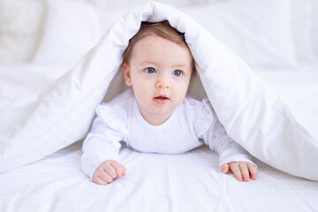 funny happy baby girl looks out from under the blanket on the bed in white clothes and smiles, a small child on a cotton bed at home woke up in the morning after sleeping