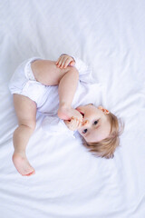 funny baby girl playing with her feet on the bed in white clothes and smiling, top view, a small child on a cotton bed at home woke up in the morning after sleeping, the concept of children's goods