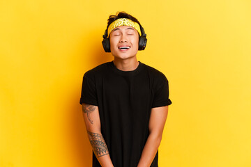 Portrait of satisfied ethnic young man with tattoo listens music via wireless headphones enjoys his favorite songs wears yellow headband and black t-shirt isolated on a yellow background. Carefree guy