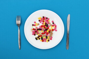 On a white plate are multi-colored pills with a knife and fork on a blue background, top view.