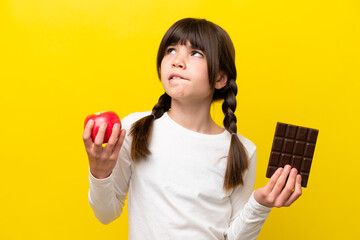 Little caucasian girl isolated on yellow background having doubts while taking a chocolate tablet...