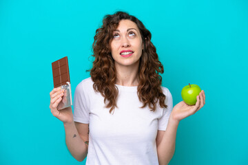 Young caucasian woman isolated on blue background having doubts while taking a chocolate tablet in...