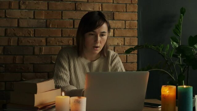 Amazed young caucasian woman using laptop to read the news via internet at home during electricity outage or blackout with lit candles. Woman shocked by the bad news touching her face in astonishment