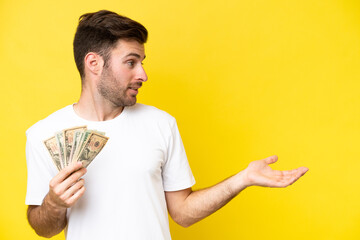 Young caucasian man taking a lot of money isolated on yellow background with surprise expression while looking side