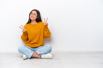 Young girl sitting on the floor isolated on white background showing victory sign with both hands