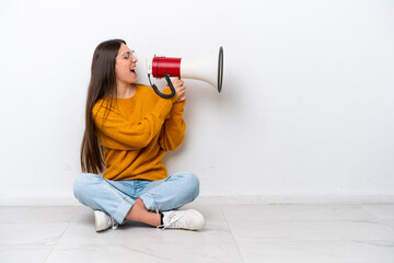 Young girl sitting on the floor isolated on white background shouting through a megaphone