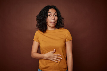 Obraz premium Young colombian curly hair woman isolated on brown background touches tummy, smiles gently, eating and satisfaction concept.