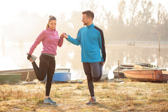 Beautiful young man and woman exercising, stretching in the early morning mist on the lake or river banks. Worming up for jogging. Healthy and active sport lifestyle