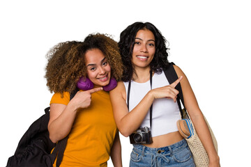 Two diverse friends on a beach vacation with vintage camera, beach bag, and travel pillow smiling and pointing aside, showing something at blank space.