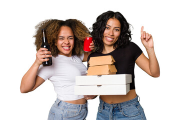 Two young women, one Latina and one with Afro hair, laugh as they hold pizzas and burgers from a...