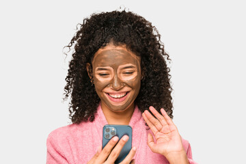 Young african american woman talking on phone while she is applying a facial mask isolated