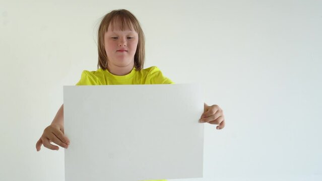 A teenage girl with blond hair in a yellow T-shirt holds a sheet of A3 paper and smiles. mock up and space for text, Down syndrome awareness concept. International Down Syndrome Day.