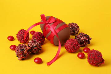 Tasty chocolate egg with red ribbon and candies on yellow background