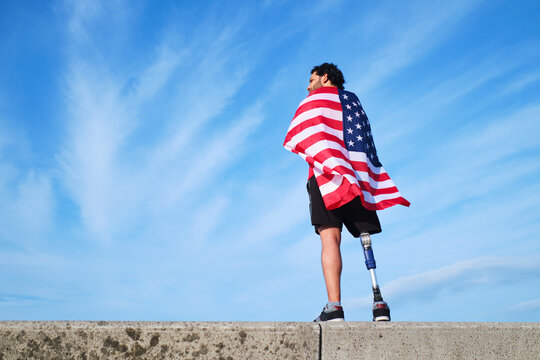 Young man with prosthetic leg standing on a retaining wall on the beach with a United States flag on his back