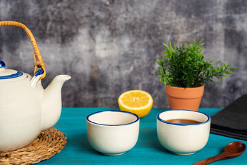 Porcelain teapot next to two cups, one with tea, with a wooden teaspoon and half a lemon, on a blue...