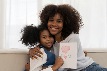 African American young mother hugging her chid daughter and holding mother day paper card at home - obrazy, fototapety, plakaty