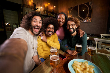 Portrait multiracial friends group looking camera at restaurant bar after work at pub happy hour. Happy friends having fun together drinking alcohol indoors. People gathered hugging for selfie.