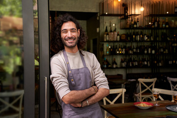 Portrait of happy young adult local small business owner looking at camera. Happy male bartender crossing his arms with positive attitude. Cheerful worker smiles while standing leaning on bar door. - obrazy, fototapety, plakaty
