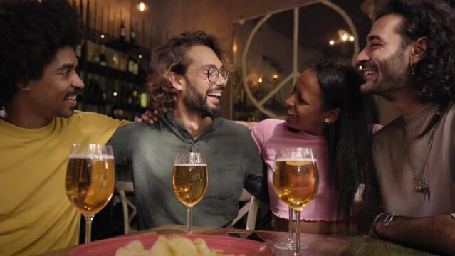 Group of multiracial colleagues gathered chatting happily at restaurant bar after work. Cheerful friends hugging having fun together drinking alcohol inside the pub. People in their free time.
