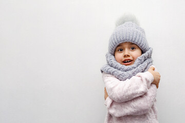 child girl in a gray hat and scarf hugs herself on a white background and smiles, grimace,...