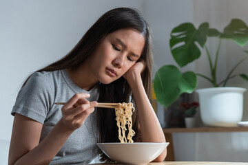 End of month. Alone depressed asian young student woman, girl using chopsticks eating instant ramen, noodles, cheap food and no money for dinner in bedroom in overtime night, late time at home. - obrazy, fototapety, plakaty