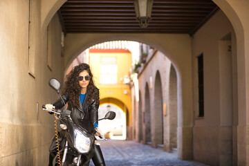 Fototapeta na wymiar Young, beautiful, brunette woman with curly hair and black leather jacket and pants, riding a black motorcycle with sexy and independent attitude. Concept beauty, motorcycles, leather, sensuality.