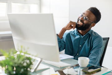 Happy black male manager sitting at office desk in front of computer, looking at screen and smiling