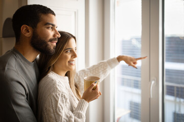 Happy young arab man hug european wife, lady pointing finger out the window and drink cup of tea