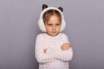 Image of little girl wearing fur earmuffs and striped shirt standing isolated over gray background, being offended, keeps arms folded, looking at cameras with pout lips.