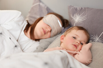 Fototapeta na wymiar Portrait of dark haired female wearing blindfold lying with her baby in bed, daughter already wake up and looking around, mommy sleeping. Happy motherhood and childhood.