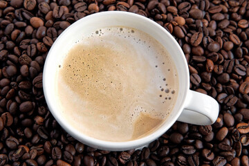 White Cup of Hot Coffee with Milk Foam on Roasted Coffee beans background. Cup of Hot Latte and Saucer. Delicious Stylish Cappuccino. Closeup. Coffee Shop. Concept for coffee product advertising.