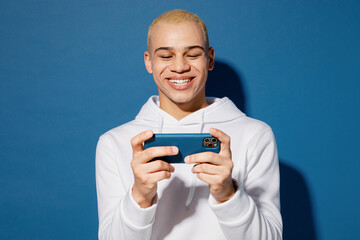 Gambling young man of African American ethnicity wear white hoody use play racing app on mobile cell phone hold gadget smartphone for pc video games isolated on plain dark royal navy blue background.